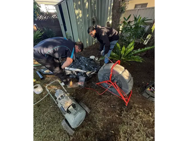 Technicians using a drain camera and drain machine to inspect an exposed sewer line in a backyard